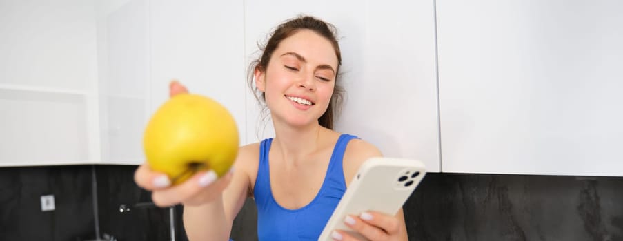 Portrait of smiling, happy fitness girl, sitting with an apple, laughing over smth on smartphone, browsing social media on mobile phone app.