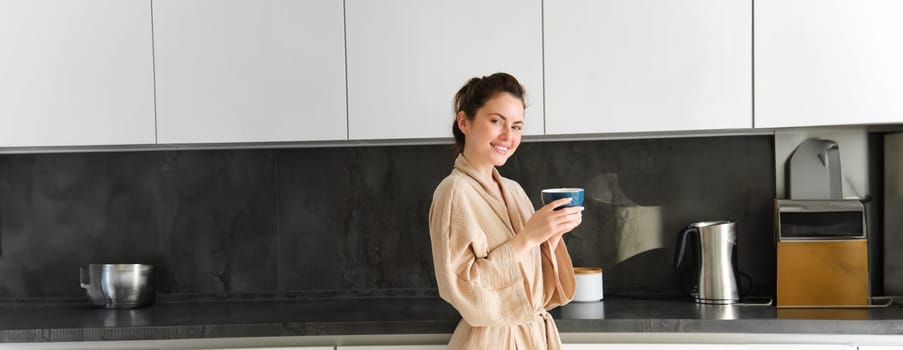 Daily routine and lifestyle. Young beautiful woman in bathrobe, standing in kitchen with cup of coffee, drinking tea, smiling and looking happy.