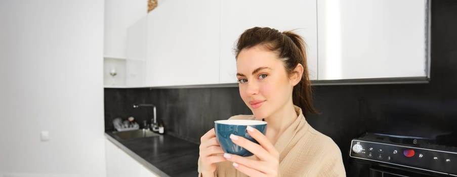 Close up portrait of attractive young woman in bathrobe, drinking coffee in the morning, holding cup of cappuccino and smiling, posing in the kitchen.