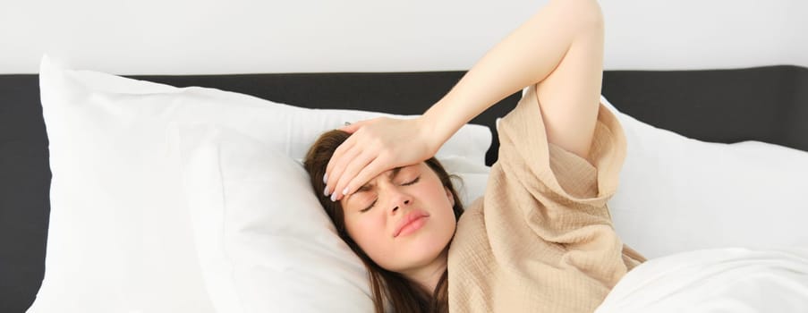 Woman lying in bed and feeling unwell, having headache, touching her head with frustrated face, has high fever or migraine. concept of health and people.