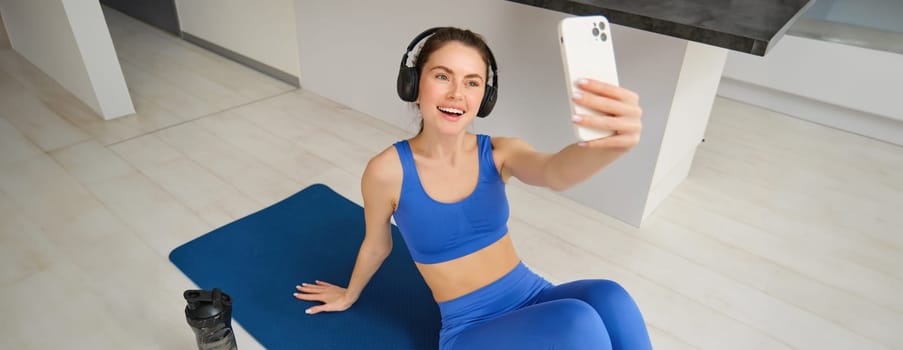 Portrait of fitness woman in wireless headphones, sits on fitness mat, doing selfies and photos during training exercises, working out at home.