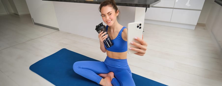 Portrait of woman taking selfie with water bottle, fitness instructor shows her exercises, doing workout from home, on rubber blue yoga mat, wearing sportswear.