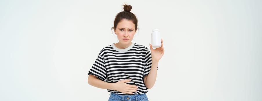 Woman takes painkillers, pills from stomach ache, menstrual pain, holds hand on belly, shows bottle with tablets, stands over white background.