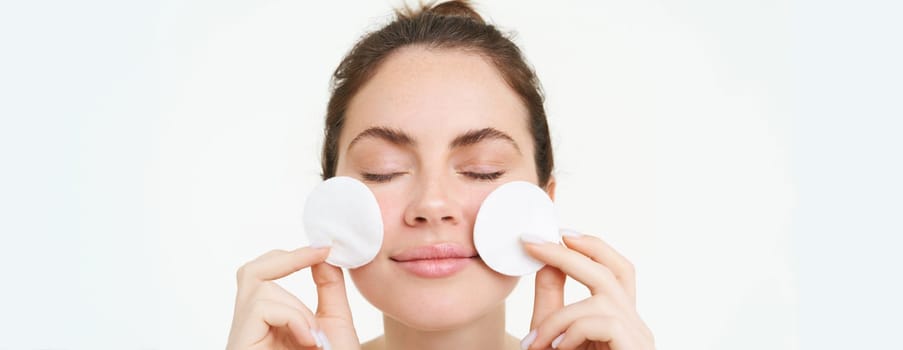 Image of young woman taking off her makeup with cotton pads, using facial cleanser, cleaning her face with skincare treatment, standing over white background.