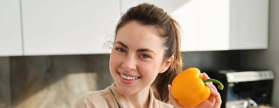 Close up portrait of beautiful woman smiling, showing yellow pepper, vegetarian girl making dinner, preparing meal with vegetables, posing in kitchen.