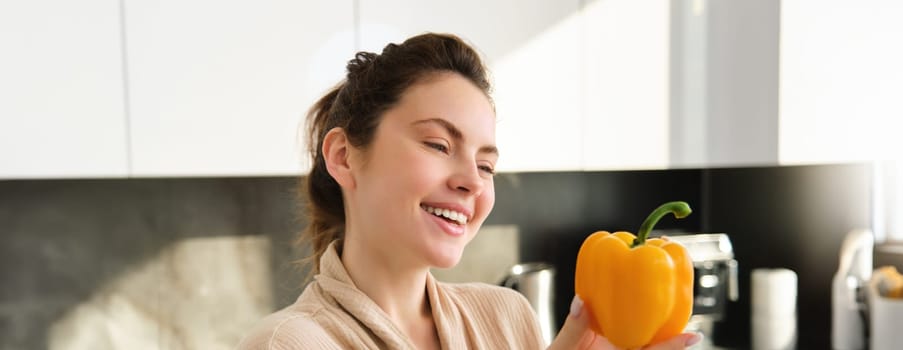 Close up portrait of beautiful woman smiling, showing yellow pepper, vegetarian girl making dinner, preparing meal with vegetables, posing in kitchen.