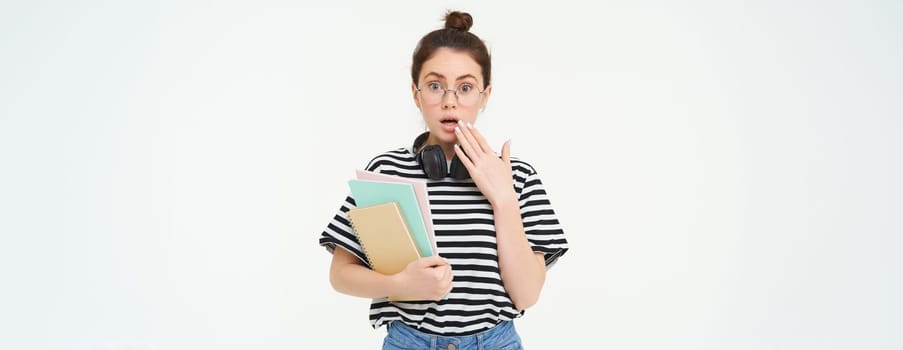 Portrait of woman in glasses, student looks surprised, gasps and looks shocked at camera, holds notebooks and study material, white background.