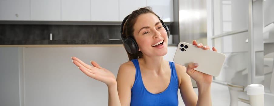 Close up portrait of happy, beautiful fitness woman, doing training exercises at home, wearing wireless headphones and singing at smartphone.