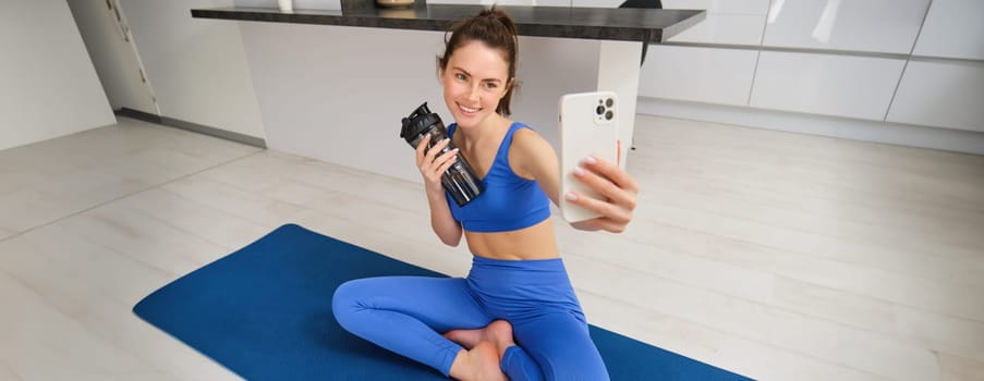 Portrait of woman doing sports, takes selfie on smartphone, fitness instructor records her exercises, stays hydrated, drinks water during training session.