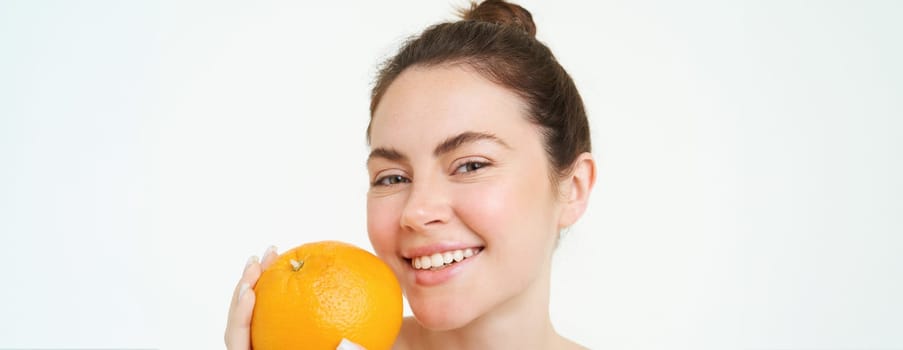 Close up portrait of smiling young woman, holding orange near face, looking happy, concept of vitamins, organic and natural skincare treatment with C serum, white background.