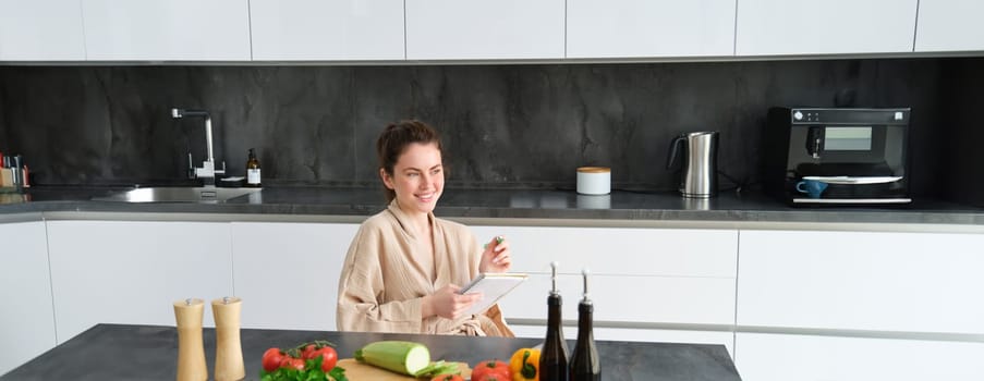 Portrait of woman writing down list of groceries, making notes in recipe, sitting in kitchen near vegetables, preparing dinner menu.