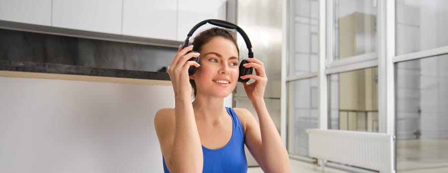 Image of young fitness girl, wearing blue tracksuit, puts on wireless headphones to workout with music, does yoga at home.