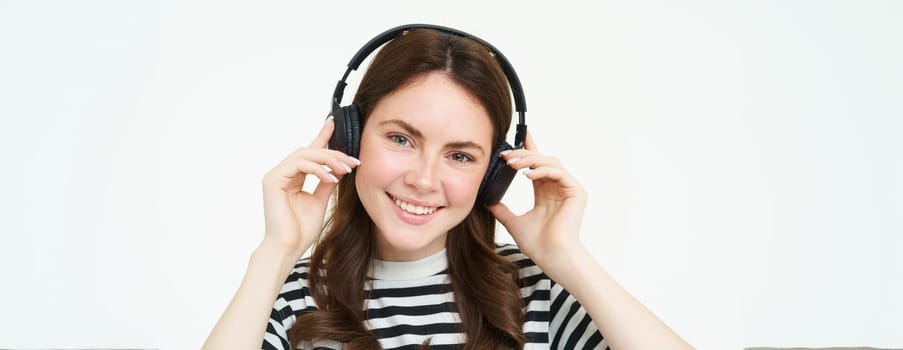 Close up portrait of cute young woman, smiling, putting on headphones, listening to music in earphones, trying on new headset, white background.