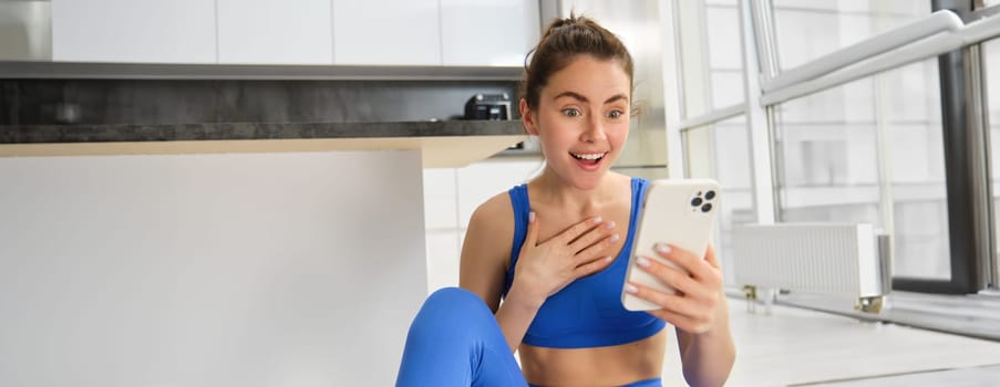 Indoor shot of surprised, happy young sportswoman looking at smartphone with smile and excitement, reading great news on phone message, sitting in blue sportsbra and leggings.
