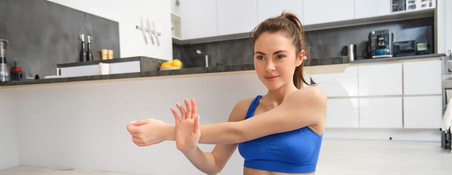 Young woman does workout from home, sits on yoga mat and stretches her hands, fitness training session in living room.