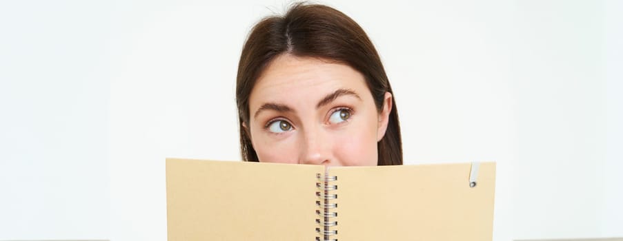 Young woman holds notebook daily planner next to her face, writing down homework, making notes, looking thoughtful, standing over white background.