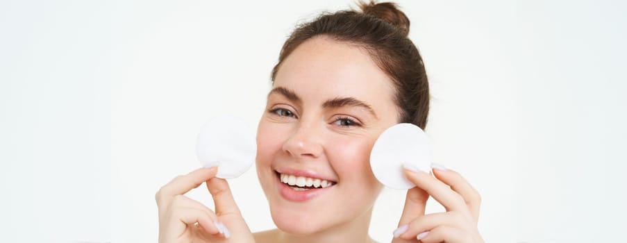 Beauty woman with clear glowing face, showing cotton cosmetic pads for makeup removal, cleansing her facial skin, standing over white background.