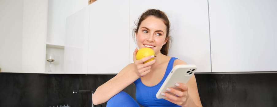 Portrait of sportswoman, girl eating and apple and looking at her social media, smartphone screen, having a snack in kitchen, wearing fitness activewear.