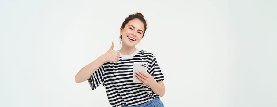 Enthusiastic girl with smartphone shows thumbs up, isolated over white background.