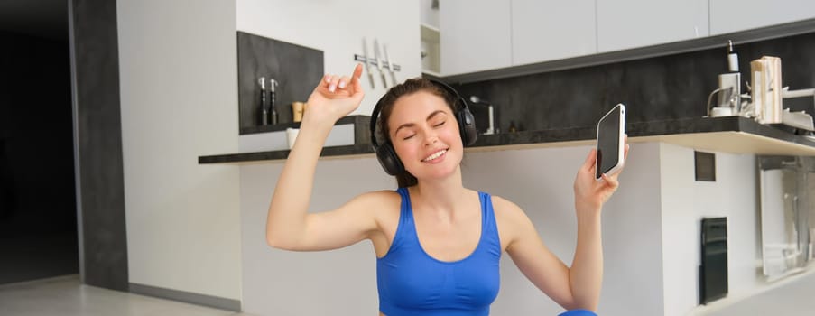 Cheerful brunette girl does fitness at home, wears wireless headphones and dances, holds smartphone, does workout indoors on floor, wears blue activewear.