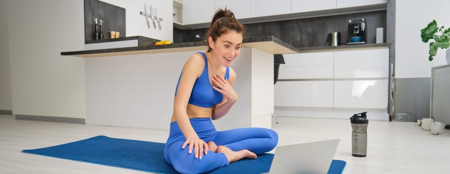 Portrait of fit and healthy, female fitness instructor sitting on floor at home, talking to laptop, teaching online class yoga or workout training.