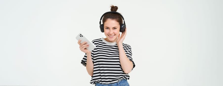 Young woman with smartphone listening to music, dancing to her favourite song in headphones, posing against white background.