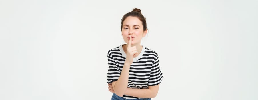 Portrait of cute girl keeps a secret, shushes, shhh gesture, presses finger to lips, gossiping, stands over white background.