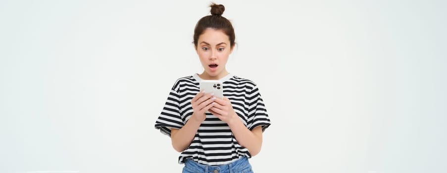 Portrait of girl looks shocked at her mobile phone, reads a message with amazed face, isolated over white background.