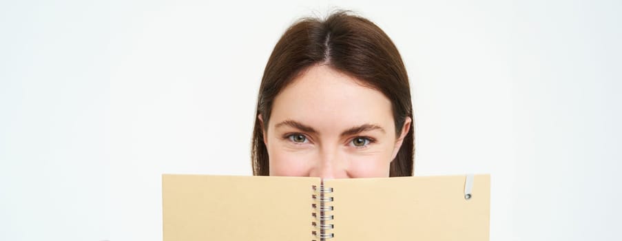 Portrait of cute young woman hides her face behind planner, holds notebook against her face and smiles, isolated over white background.
