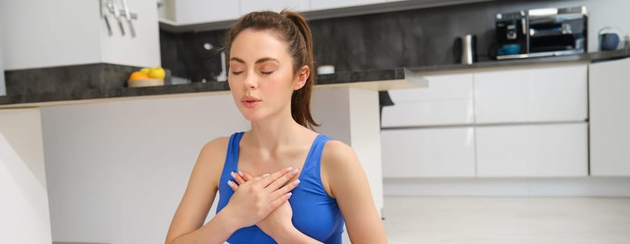 Image of calm and relaxed woman meditating, doing breathing practices, holding hands on chest during yoga session at home. Sport and mindfulness concept