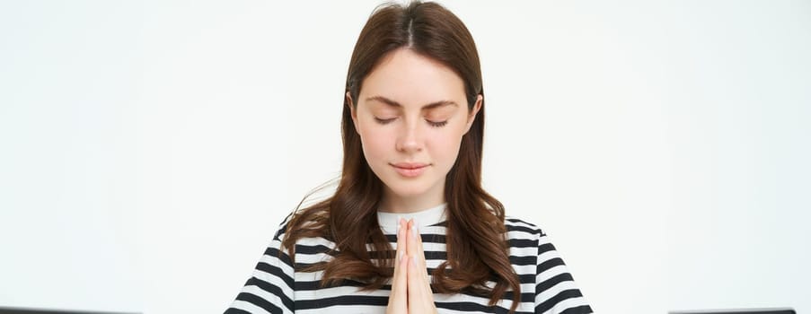 Portrait of woman showing namaste gesture, holding palms clenched together near chest, standing isolated over white background.