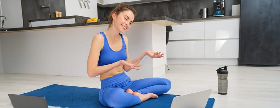 Image of young smiling woman, fitness instructor sitting on yoga mat, talking to client via online video chat on laptop, teaching aerobics, workout training.