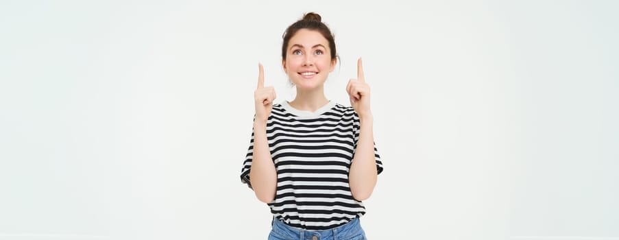Portrait of excited, smiling young woman, looking and pointing up with happy, emotional face, white background.