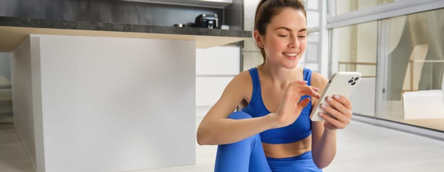 Image of young fit woman wearing sportbra and blue leggings, using smartphone app, does workout from home, sits on floor and follows online training video.