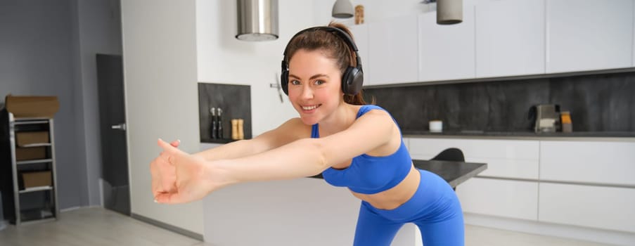 Portrait of beautiful, fit woman in blue leggings and sportsbra, stretching her arms, doing fitness workout, aerobics exercises at home, listening music in wireless headphones.
