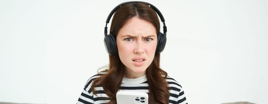 Image of puzzled young woman in headphones, holds smartphone, frowning and looking confused at camera, standing over white background.
