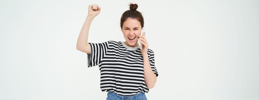 Enthusiastic girl chanting, answers phone call and looks excited, triumphing over great news, stands over white background.
