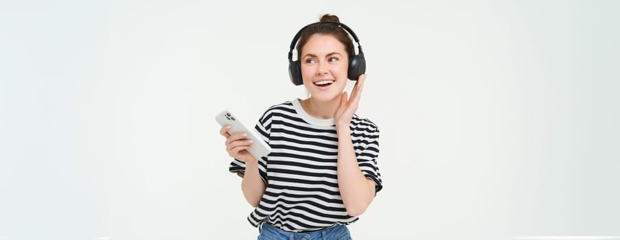 Portrait of happy woman with smartphone changes song on mobile phone streaming app, listens music in headphones, dancing against white background.