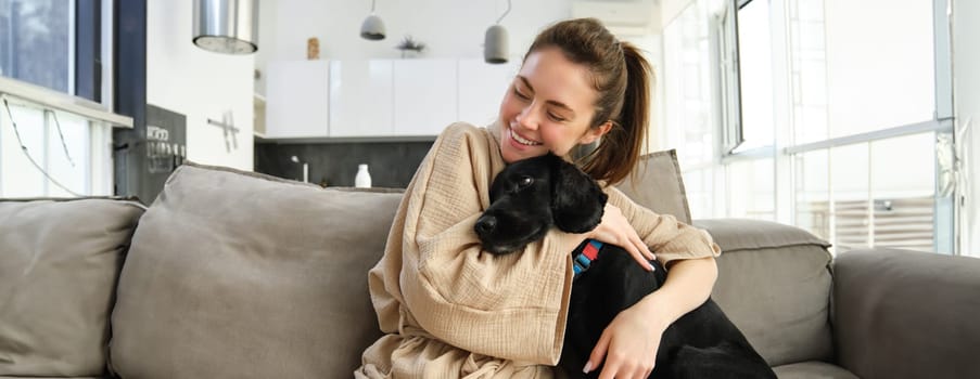 Animals and lifestyle concept. Happy young woman in bathrobe, hugs her dog on sofa, cuddle puppy and smiling.