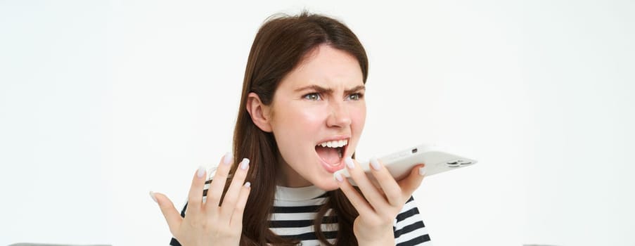 Portrait of angry woman shouting at smartphone speakerphone, recording voice message with annoyed face expression, screaming at phone, white background.