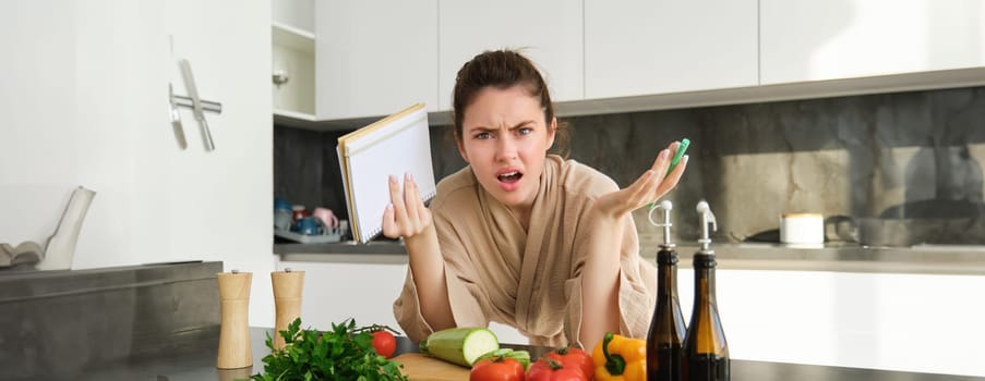 Portrait of woman with angry face, standing near vegetables and looking frustrated, holding notebook, annoyed while cooking meal. Lifestyle and people concept