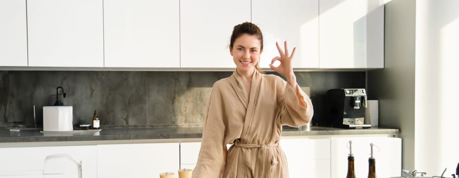 Portrait of smiling brunette woman cooking healthy dinner, showing okay sign, wife standing in bathrobe in kitchen, preparing food, chopping vegetables on board.