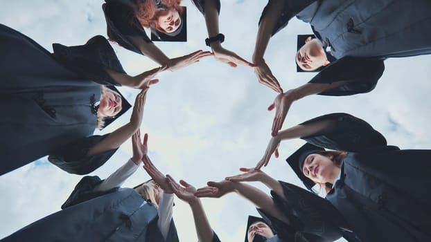 Graduate students in black robes and caps make a circle shape from their hands