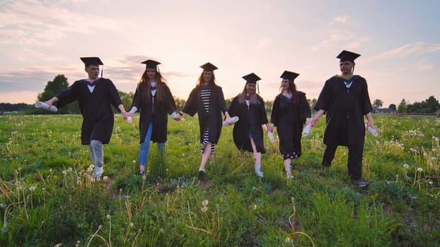 Six graduates in robes walking through a meadow in the evening