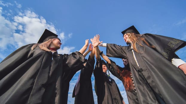 Team of college or university students celebrating graduation. Group of happy successful graduates in academic hats and robes standing in circle and putting their hands together