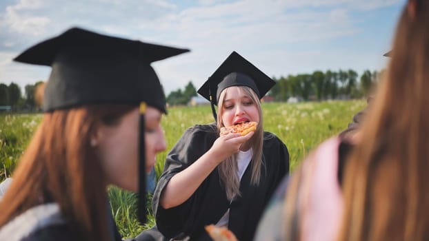 Graduates in black suits eating pizza in a city meadow