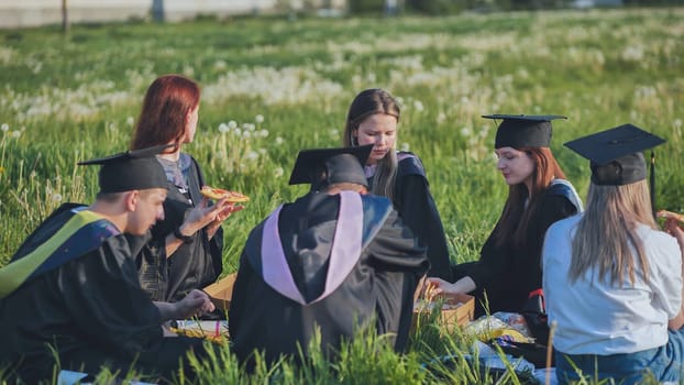 Graduates in black suits eating pizza in a city meadow