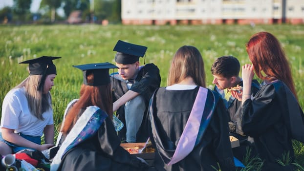 Graduates in black suits eating pizza in a city meadow