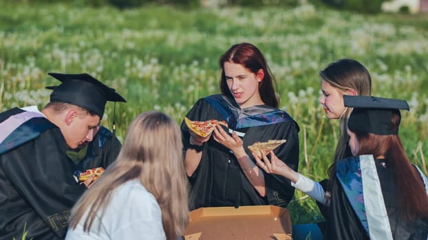 Graduates in black suits eating pizza in a city meadow