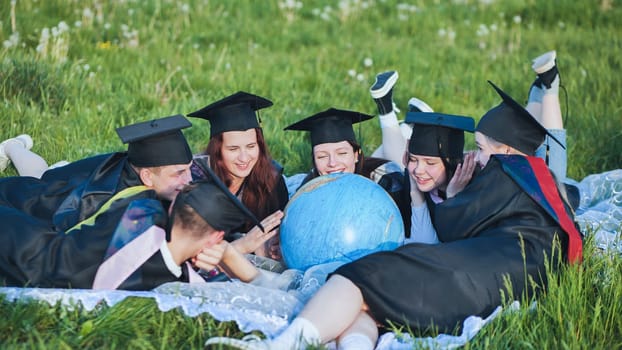 Graduates in black robes looking at a globe lying on the grass